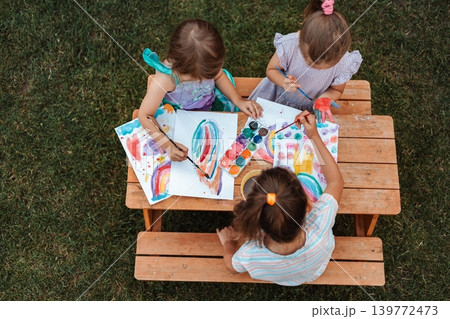 Three little girls painting with watercolors at a wooden table in a garden 139772473