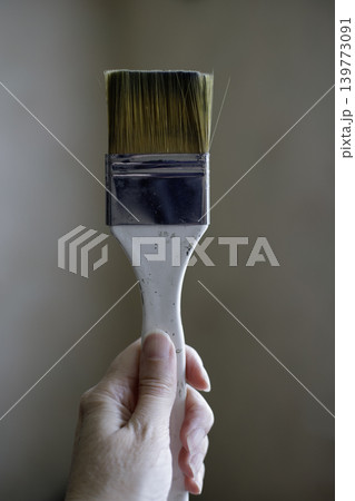 Hand holding clean bristle paint brush vertically on a blurred grey wall background, repair tools for interior house decorating close up 139773091