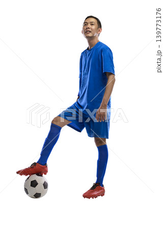 A young Indonesian southeast asian male athlete in a full blue soccer kit stands confidently with one foot resting on a soccer ball, isolated against a white background 139773176