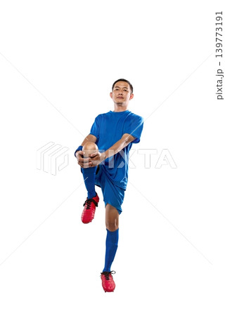 Professional Indonesian southeast asian soccer player in blue uniform performing a knee-to-chest leg stretch, isolated on a white background. Ideal for fitness and sports marketing Professional Indonesian southeast asian soccer player in blue uniform performing a knee-to-chest leg stretch, isolated on a white background. Ideal for fitness and sports marketing 139773191