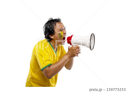 An excited Indonesian southeast asian male soccer fan wearing a yellow and green jersey shouting into a megaphone and cheering with his fist raised, isolated on white background An excited Indonesian southeast asian male soccer fan wearing a yellow and green jersey shouting into a megaphone and cheering with his fist raised, isolated on white background 139773253