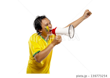 An excited Indonesian southeast asian male soccer fan wearing a yellow and green jersey shouting into a megaphone and cheering with his fist raised, isolated on white background 139773254