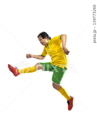 Full length shot of a professional Indonesian southeast asian male soccer player in a yellow and green uniform performing a dynamic jumping kick, isolated on white background. Full length shot of a professional Indonesian southeast asian male soccer player in a yellow and green uniform performing a dynamic jumping kick, isolated on white background. 139773260