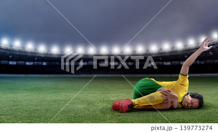 Professional Indonesian southeast asian male soccer player in yellow and green uniform lies on the stadium grass in pain, clutching her injured knee during a match under bright floodlights 139773274
