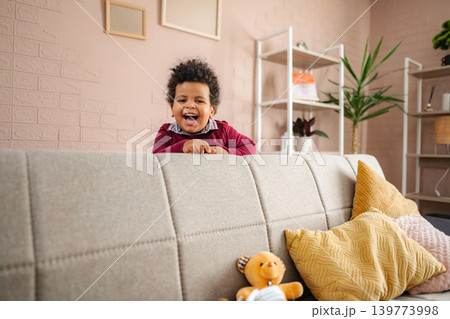 Happy preschool age boy playing playful hiding behind sofa Happy preschool age boy playing playful hiding behind sofa 139773998
