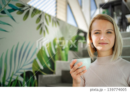 Woman holding coffee cup, smiling indoors, tropical mural 139774005