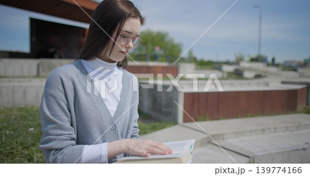 A hand-held shot of a young female student in casual clothes and glasses reading a book while flipping through pages while sitting in an urban park. 139774166