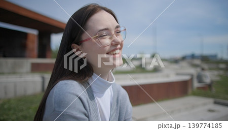 A handheld shot of a young female student in casual clothes sitting on a concrete bench in an urban park, enjoying a sunny summer day. A handheld shot of a young female student in casual clothes sitting on a concrete bench in an urban park, enjoying a sunny summer day. 139774185
