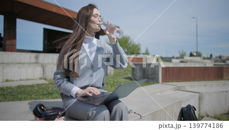 A hand-held shot of a young female student in casual clothes wearing glasses sitting on a concrete bench in an urban park on a sunny summer day, working on a laptop and typing on a keyboard. A hand-held shot of a young female student in casual clothes wearing glasses sitting on a concrete bench in an urban park on a sunny summer day, working on a laptop and typing on a keyboard. 139774186