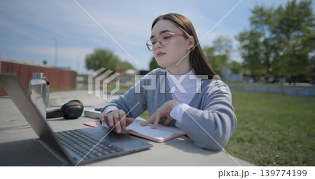 A hand-held shot of a young female student in casual clothes and glasses using a laptop and taking notes with a pen in a notebook in an urban park. 139774199