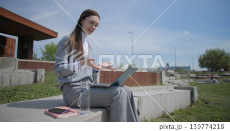 A hand-held shot of a young female student in casual clothes and glasses conducting a video call or conference on a laptop while sitting in an urban park A hand-held shot of a young female student in casual clothes and glasses conducting a video call or conference on a laptop while sitting in an urban park 139774218
