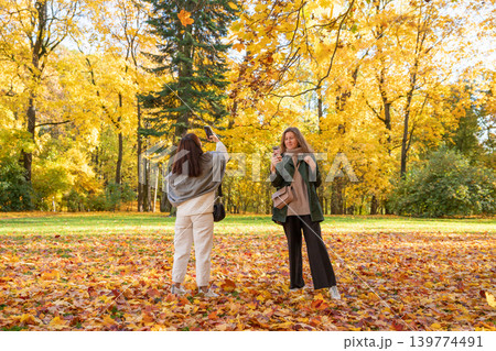 Woman taking photo of friend with smartphone in autumn park. Two women enjoying fall season outdoors. Golden leaf foliage in forest during weekend leisure. Friends spending time together. 139774491