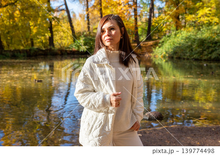 Woman standing in autumn park near lake. Beautiful middle aged lady wearing white jacket during fall season. Outdoor leisure and calm mood in nature for lifestyle blog or concept. 139774498