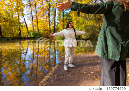 Woman in white jacket standing with open arms near pond in autumn park. Happy girl greeting friend in forest. Seasonal weekend activity, recreation in nature and friendship concept. Woman in white jacket standing with open arms near pond in autumn park. Happy girl greeting friend in forest. Seasonal weekend activity, recreation in nature and friendship concept. 139774499