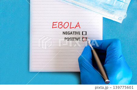Ebola virus words symbol. Concept word Ebola positive on beautiful white note. Beautiful blue background. Doctor hand in blue medical glove. Medical mask. Medical Ebola virus concept. Copy space. Ebola virus words symbol. Concept word Ebola positive on beautiful white note. Beautiful blue background. Doctor hand in blue medical glove. Medical mask. Medical Ebola virus concept. Copy space. 139775601