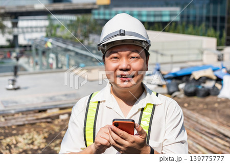 Professional worker checks mobile application near a construction project with modern infrastructure, protective equipment, and urban development visible in the background. 139775777