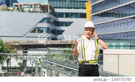 Construction supervisor talking on smartphone while coordinating workers and managing operations at modern construction site. 139775797