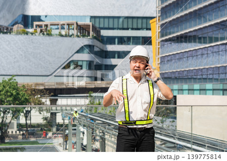 Serious construction supervisor talking on smartphone with worried expression while coordinating urgent issues at construction site. Serious construction supervisor talking on smartphone with worried expression while coordinating urgent issues at construction site. 139775814