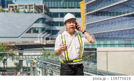 Serious construction supervisor talking on smartphone with worried expression while coordinating urgent issues at construction site. Serious construction supervisor talking on smartphone with worried expression while coordinating urgent issues at construction site. 139775819