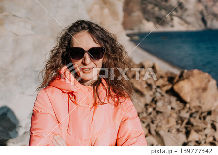 Woman coastline jacket stands on a rocky beach wearing a bright peach winter coat and sunglasses during a sunny day 139777342