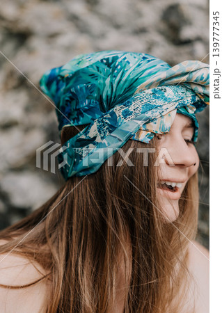 Woman headscarf smiling outdoors as she wears a vibrant blue tropical patterned wrap against a rocky background Woman headscarf smiling outdoors as she wears a vibrant blue tropical patterned wrap against a rocky background 139777345