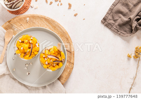 Overhead view of two glass jars filled with fresh mango chunks and coconut flakes on a round wooden board and grey plate surrounded by rustic linens and copy space for a bright healthy concept Overhead view of two glass jars filled with fresh mango chunks and coconut flakes on a round wooden board and grey plate surrounded by rustic linens and copy space for a bright healthy concept 139777648