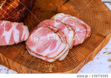 Sliced cured pork meat is displayed on wooden cutting board with whole piece nearby in kitchen setting. Sliced cured pork meat is displayed on wooden cutting board with whole piece nearby in kitchen setting. 139778395