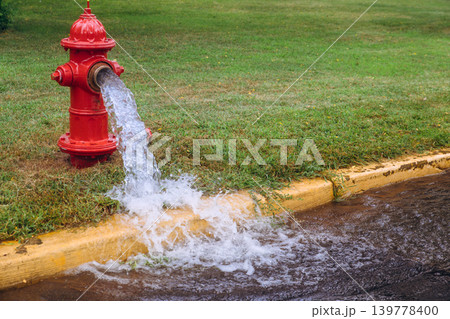 Water flows from leaking red fire hydrant into street while grass grows nearby in suburban neighborhood. Water flows from leaking red fire hydrant into street while grass grows nearby in suburban neighborhood. 139778400