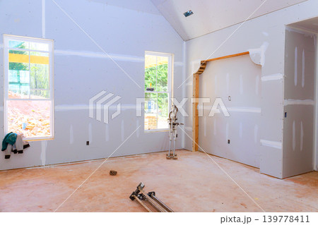 Workers are installing drywall in room with light coming through windows while construction continues outside. Workers are installing drywall in room with light coming through windows while construction continues outside. 139778411