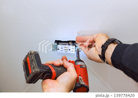 Worker is fixing an electrical outlet on celling uses screwdriver to secure outlet in place. 139778420