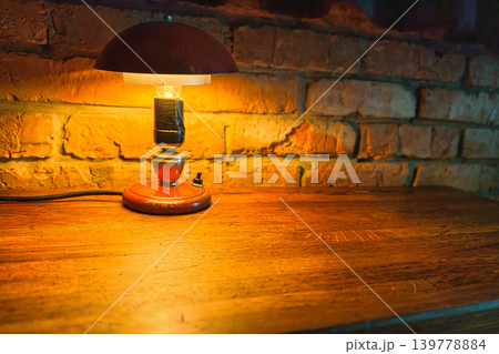 A lamp sits on a wooden table with a warm glow. The lamp is next to a brick wall in a quiet space during evening hours A lamp sits on a wooden table with a warm glow. The lamp is next to a brick wall in a quiet space during evening hours 139778884