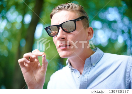 Sunglasses, man and smoking weed outdoor at event, celebration and music festival alone. Cigarette, marijuana and face of young person at party, carnival and concert in nature with cannabis joint 139780157