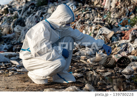 Environmentalist picks up dirty plastic from a polluted site while wearing a protective suit and gloves. The scene shows a landfill with waste Environmentalist picks up dirty plastic from a polluted site while wearing a protective suit and gloves. The scene shows a landfill with waste 139780228