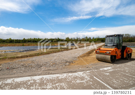 Asphalt paver is actively moving along a construction site near a water body under bright sky with few clouds in view 139780231