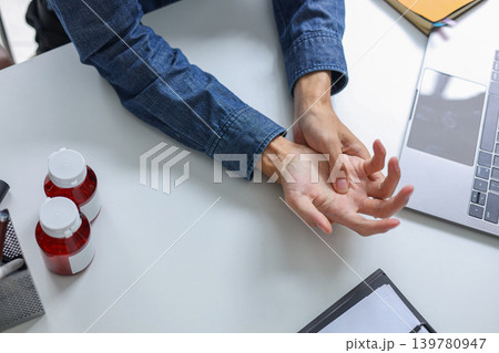 Employee suffering from wrist or hand pain, carpal tunnel syndrome, arthritis while working on laptop with medicine bottle at desk. 139780947