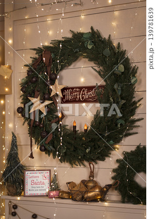 Large Christmas wreath on wooden wall with garland lights and festive signage in cozy home Large Christmas wreath on wooden wall with garland lights and festive signage in cozy home 139781639