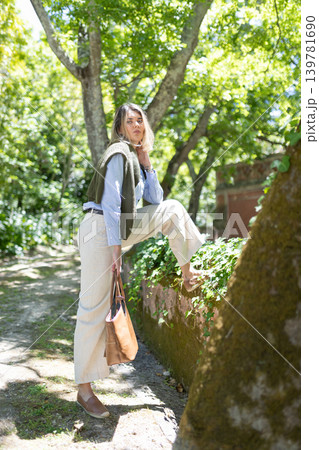 Woman stands on stone wall in green park while holding tan bag and looking at camera during bright sunny day 139781690