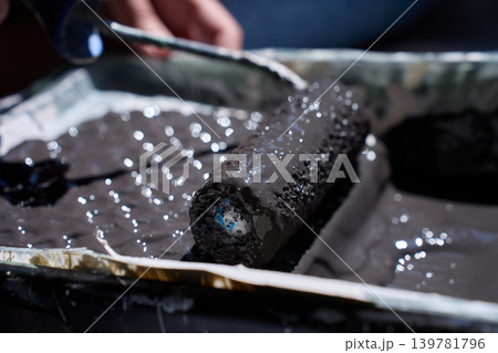 Close up of paint roller resting in tray with dark paint and hand in background 139781796