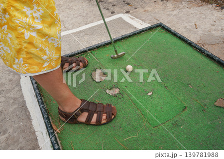 Young man playing minigolf on old, worn hotel resort green field, closeup to his feet in sandals, metal club and ball Young man playing minigolf on old, worn hotel resort green field, closeup to his feet in sandals, metal club and ball 139781988