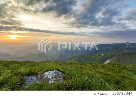 Wide summer mountain view at sunrise. Glowing orange sun raising in blue cloudy sky over green grassy hill with big rock and distant mountain range covered with morning mist. Beauty of nature concept. Wide summer mountain view at sunrise. Glowing orange sun raising in blue cloudy sky over green grassy hill with big rock and distant mountain range covered with morning mist. Beauty of nature concept. 139782344