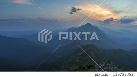 Fantastic sunrise or sunset over green mountain ridge covered with dense blue fog. Bright orange sun raising in soft cloudy sky over distant horizon. Beauty of nature, tourism and traveling concept. 139782352