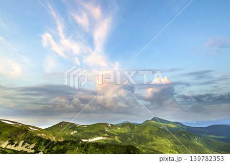 Wide panorama of lit by morning sun green valley, hills covered with forest and distant misty mountains with patches of snow under cloudy windy sky. Beauty of nature, tourism and traveling concept. 139782353