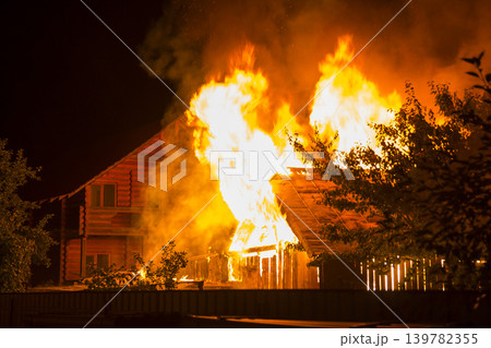 Burning wooden house at night. Bright orange flames and dense smoke from under the tiled roof on dark sky, trees silhouettes and residential neighbor cottage background. Disaster and danger concept. Burning wooden house at night. Bright orange flames and dense smoke from under the tiled roof on dark sky, trees silhouettes and residential neighbor cottage background. Disaster and danger concept. 139782355