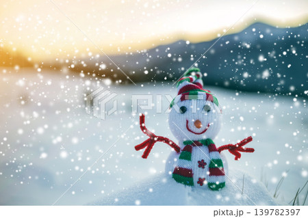 Small funny toy baby snowman in knitted hat and scarf in deep snow outdoor on blurred mountains landscape and falling big snowflakes background. Happy New Year and Merry Christmas greeting card theme. Small funny toy baby snowman in knitted hat and scarf in deep snow outdoor on blurred mountains landscape and falling big snowflakes background. Happy New Year and Merry Christmas greeting card theme. 139782397