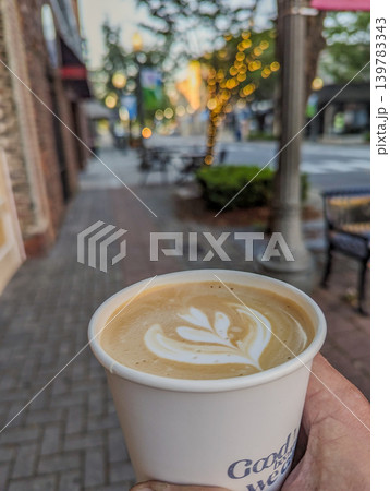 Hand Holding Takeaway Coffee Cup with Latte Art on Urban Street with Soft Bokeh Lights Background 139783343