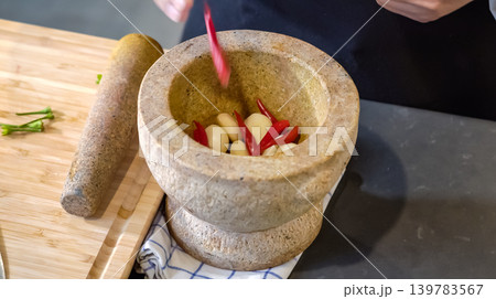 A chief puts chilies and garlics in the stone mortar and pounding with stone pestle on the counter. Preparing food ingredients. 139783567