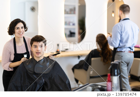Adult woman hairdresser gives haircut to young guy Adult woman hairdresser gives haircut to young guy 139785474