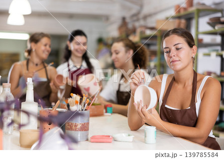 Young woman sits near table and decorates clay ceramic craft vase, plate Young woman sits near table and decorates clay ceramic craft vase, plate 139785584