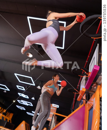Sportive adult female in active wear throwing ball in basketball basket while jumping on trampolines in entertainment center Sportive adult female in active wear throwing ball in basketball basket while jumping on trampolines in entertainment center 139785836