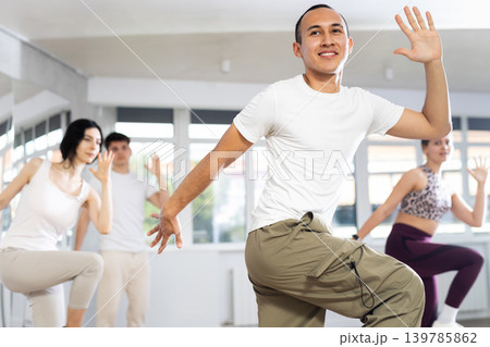 Positive guy and group of young active sport people practicing vogue dancing movements while training twist or charleston in dance hall 139785862
